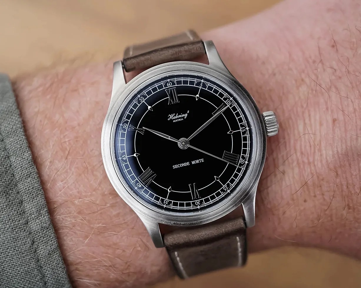 Wristwatch with black dial, silver case, Roman numerals, and brown leather strap worn on a wrist.
