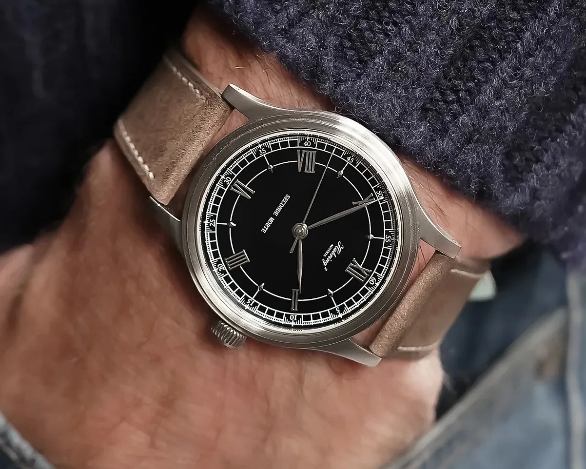 Silver wristwatch with black dial, Roman numerals, and tan leather strap worn on a wrist.