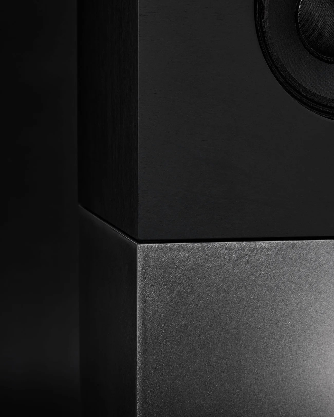 Close-up of a black speaker cabinet with a textured dark gray metal base against a black background.