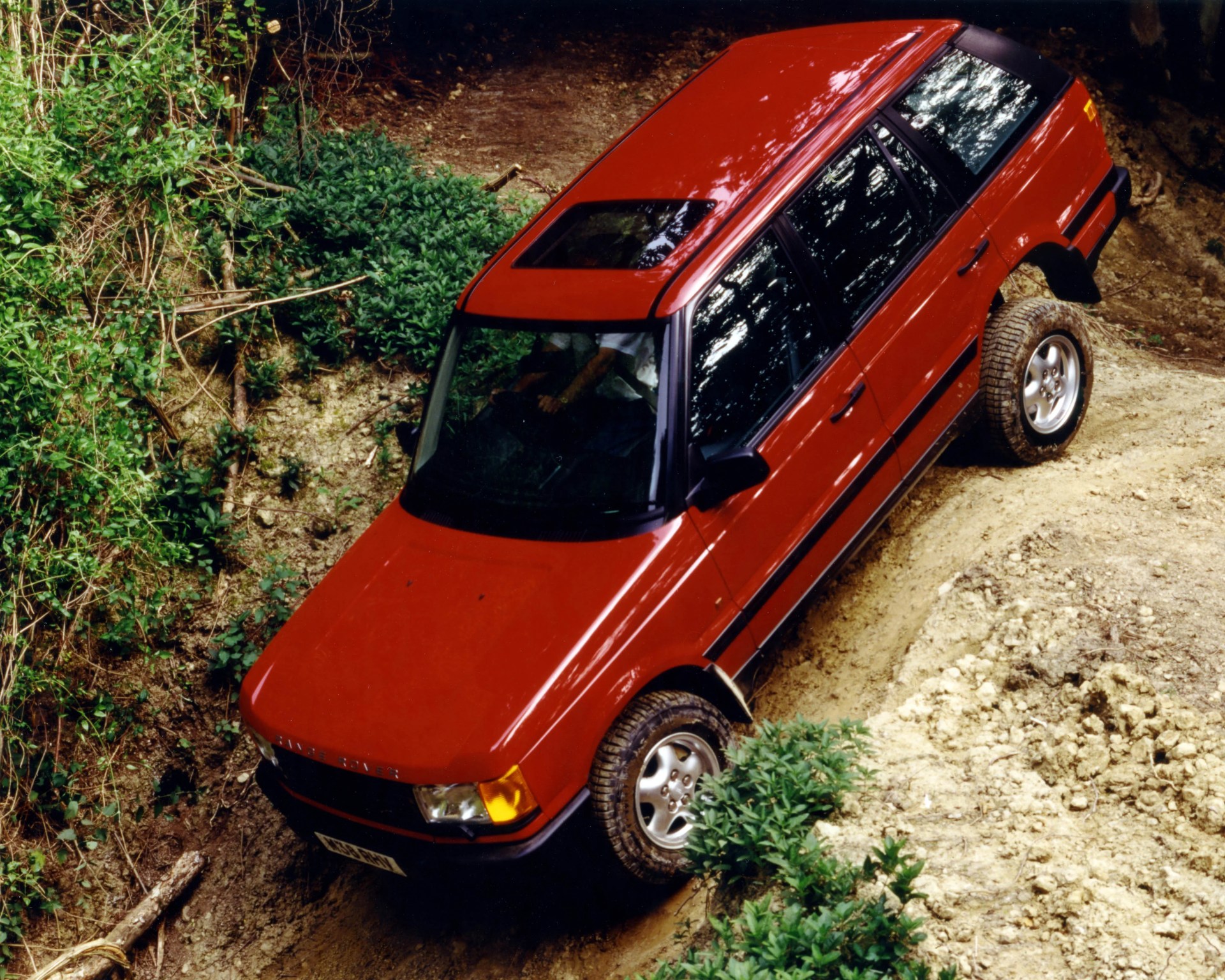Red Range Rover SUV driving on a rough dirt trail surrounded by greenery.
