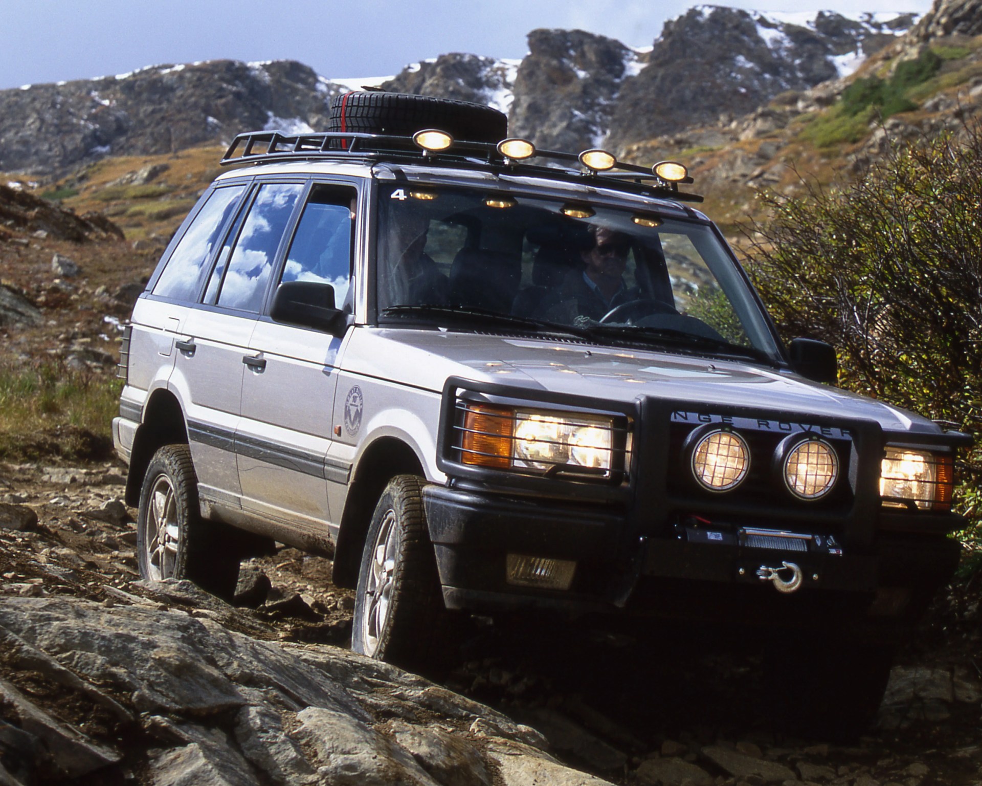 Silver Land Rover SUV with roof rack and spare tire driving on rocky mountain terrain.