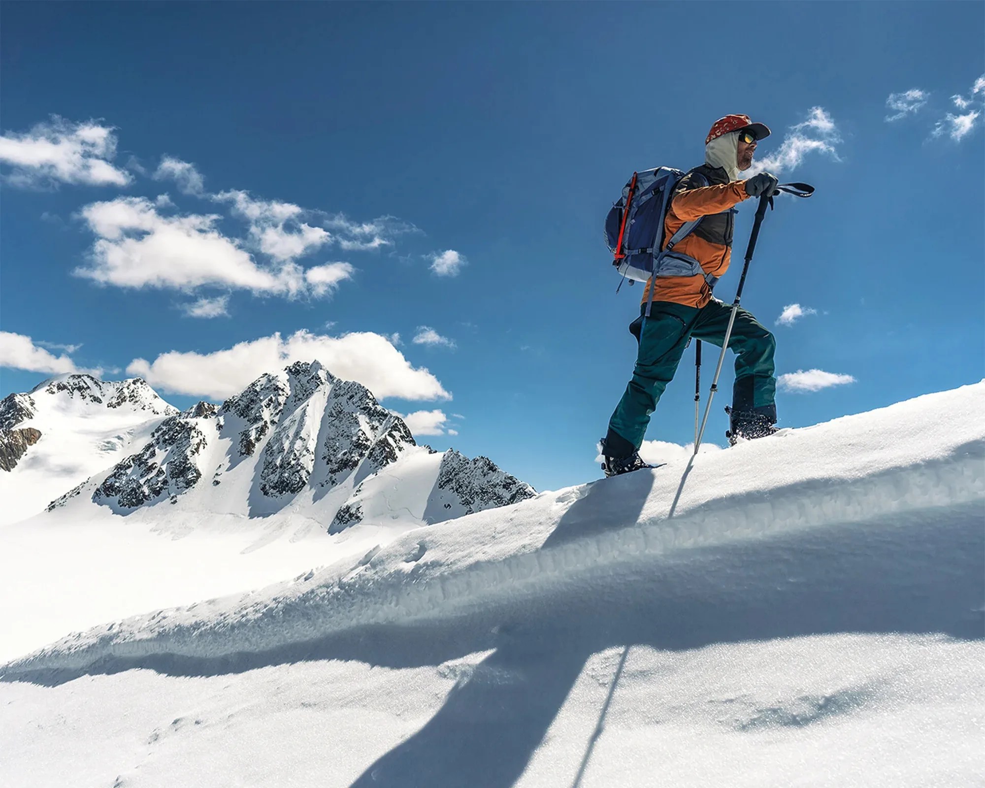 Person wearing orange jacket and green pants hiking with poles on snowy mountain under blue sky.