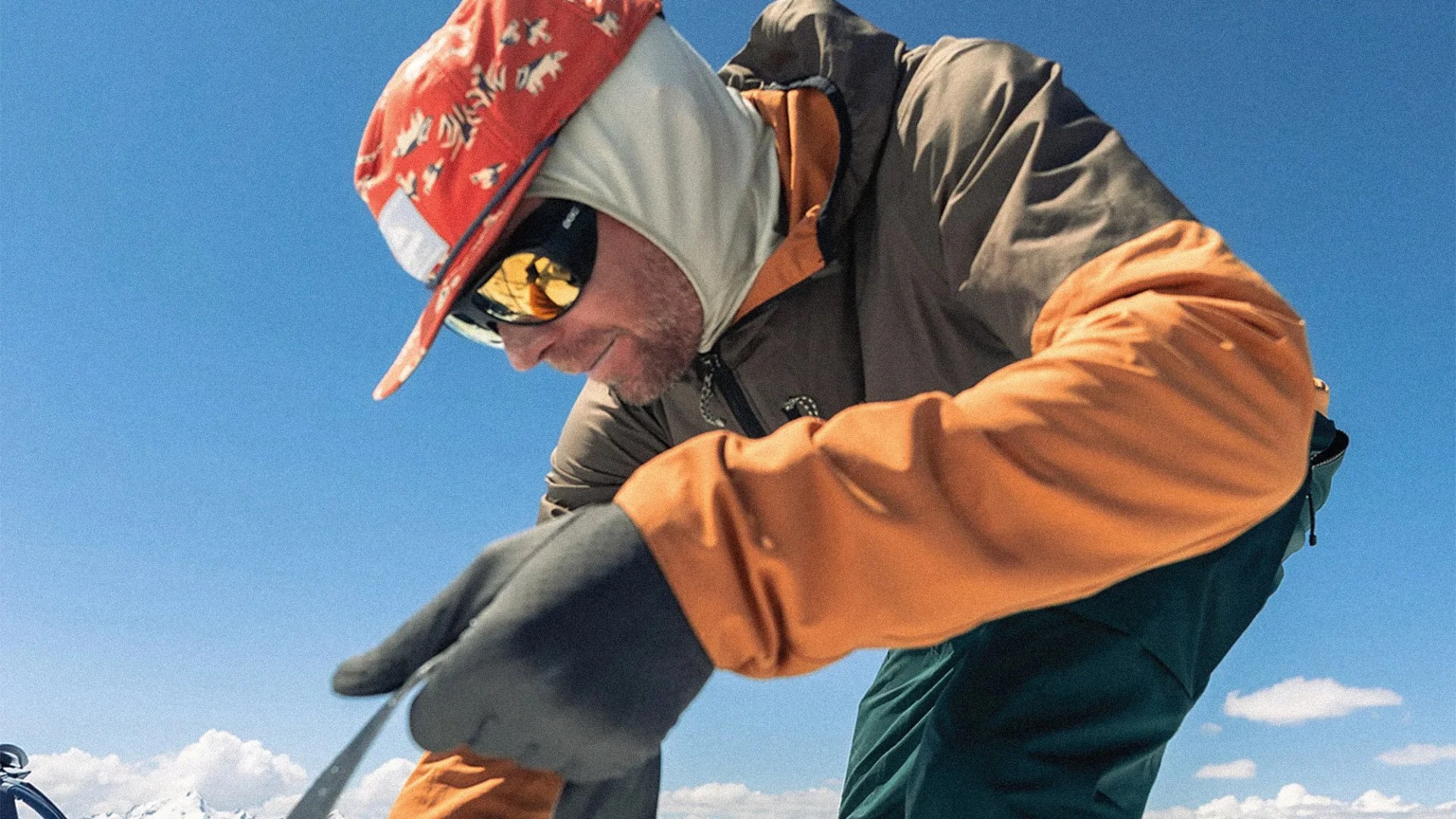 Man wearing orange and brown jacket, red patterned cap, and reflective sunglasses against a clear blue sky.