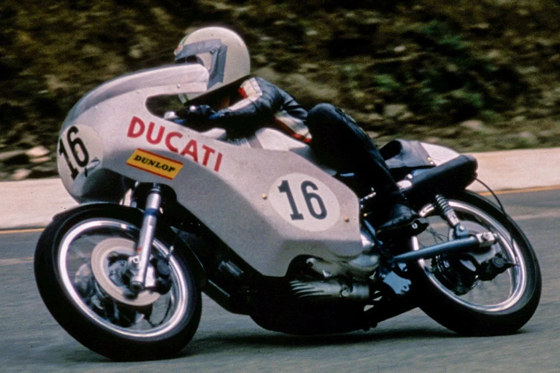 Rider in white helmet on vintage white Ducati racing motorcycle number 16 leaning into a turn on a paved road.