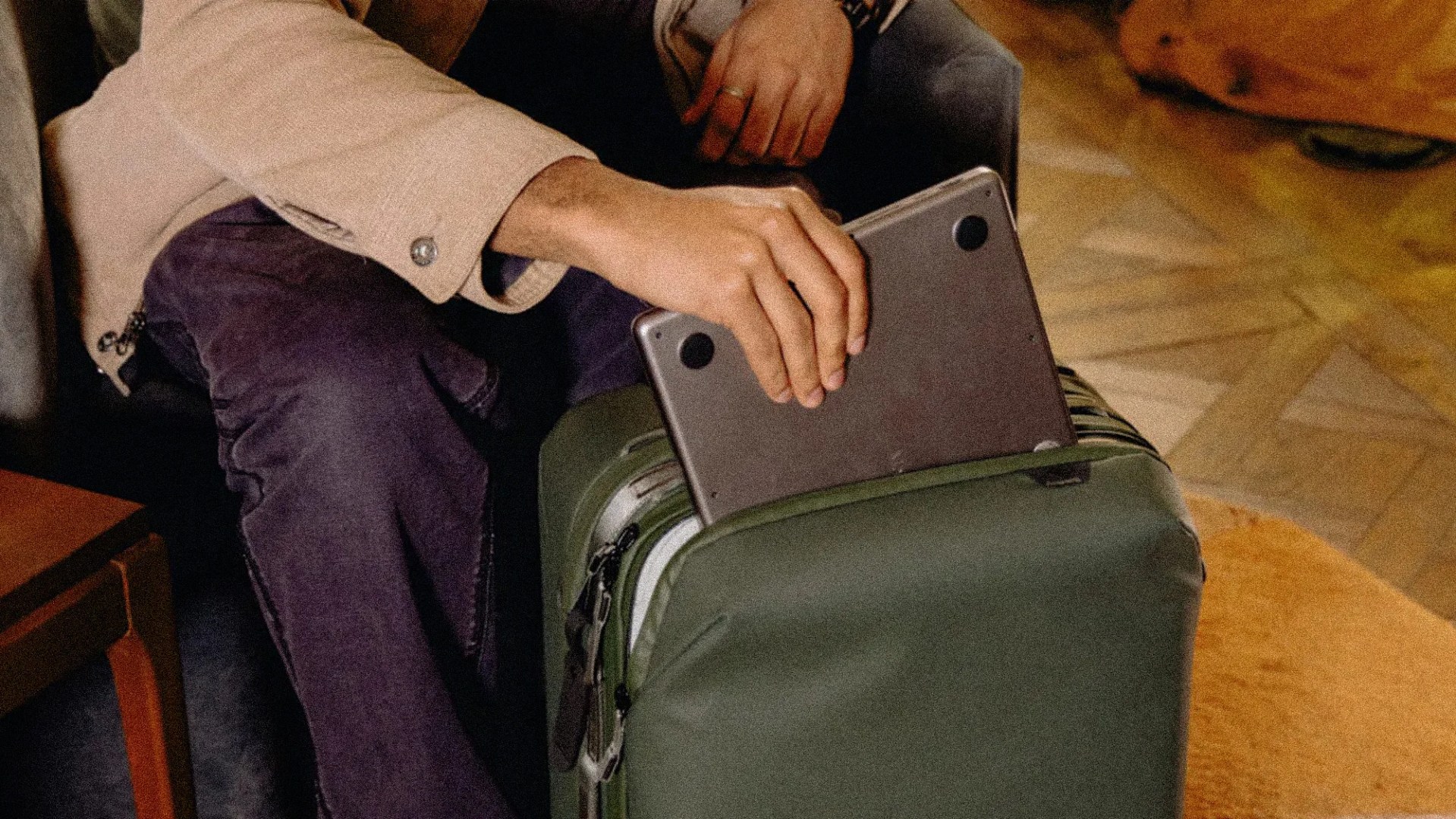 Person placing a silver laptop into the front pocket of a green suitcase.