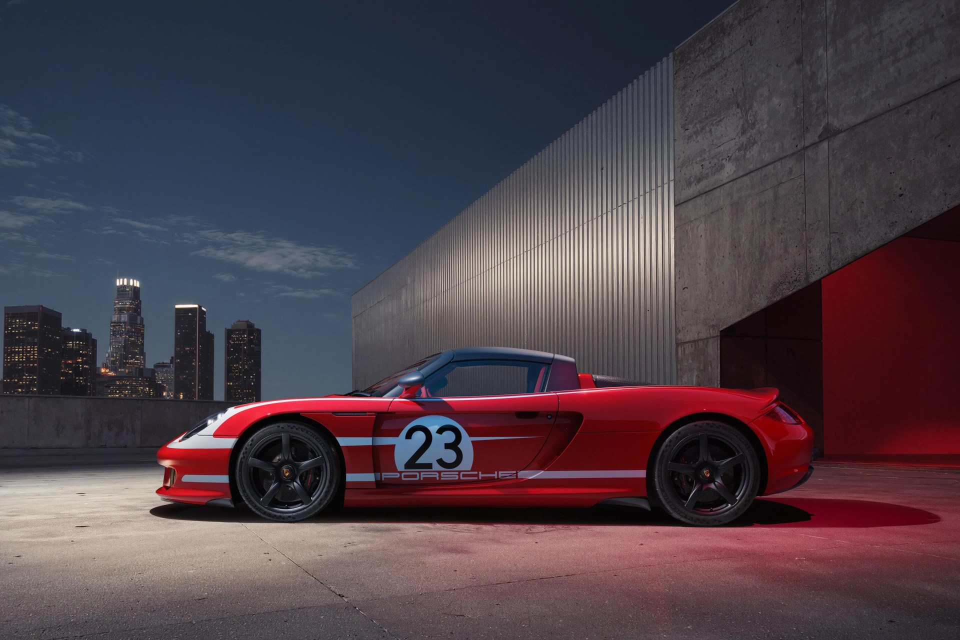 Red Porsche convertible with white racing stripes and number 23 on the side, parked at night by a concrete and metal building.
