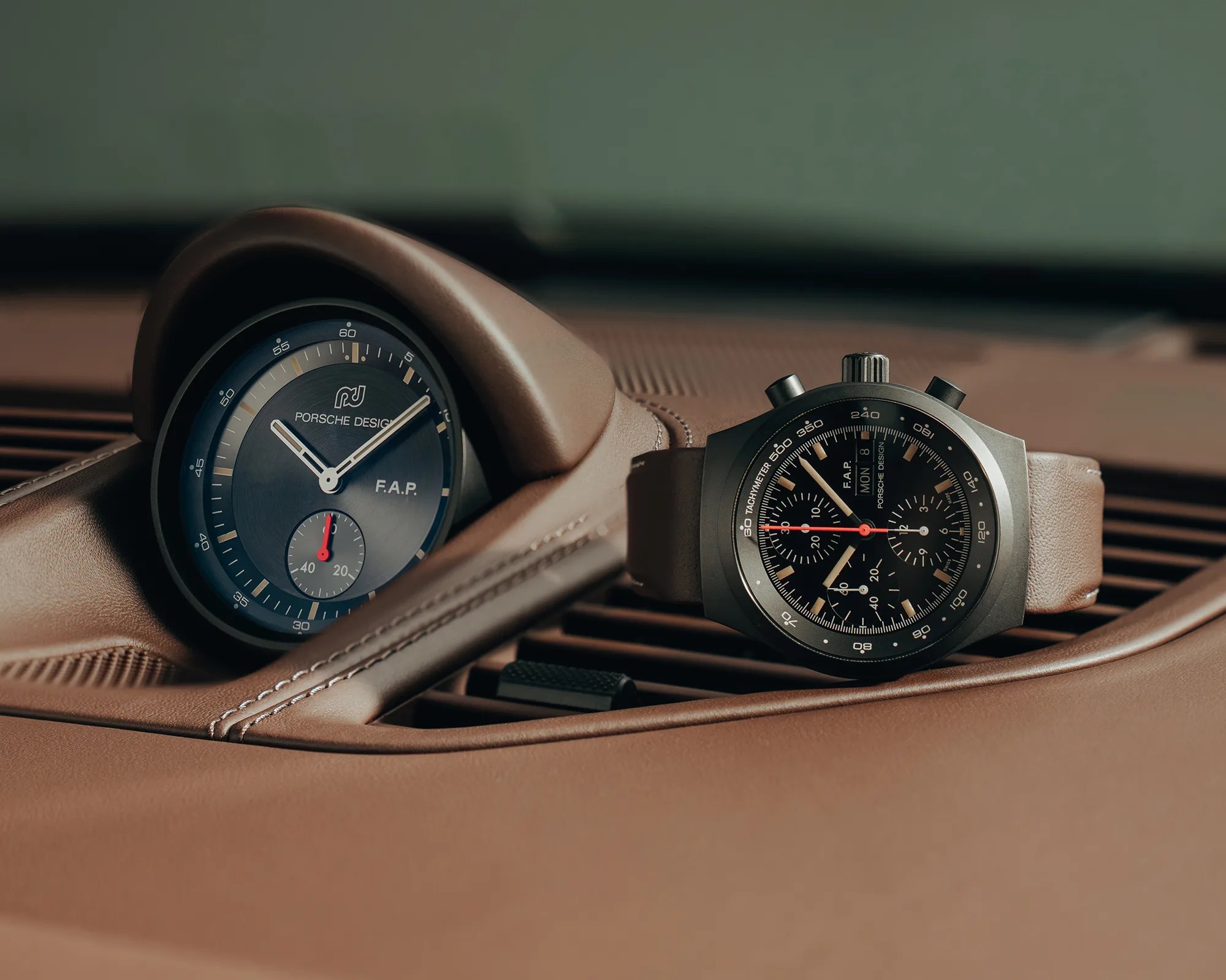 Black Porsche Design chronograph watch with brown leather strap next to a Porsche Design dashboard clock in a brown leather car interior.