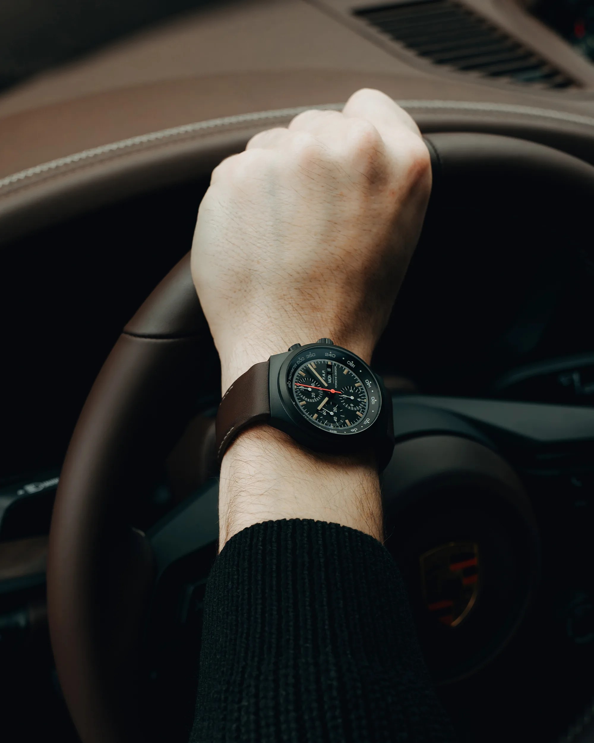 Black chronograph wristwatch with brown leather strap worn on a wrist gripping a brown leather steering wheel.