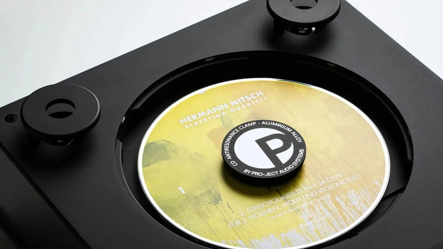 Close-up of a black turntable with a yellow CD labeled "Hermann Nitsch Albertina Quartett" secured by a Pro-Ject Audio Systems anti-resonance clamp.