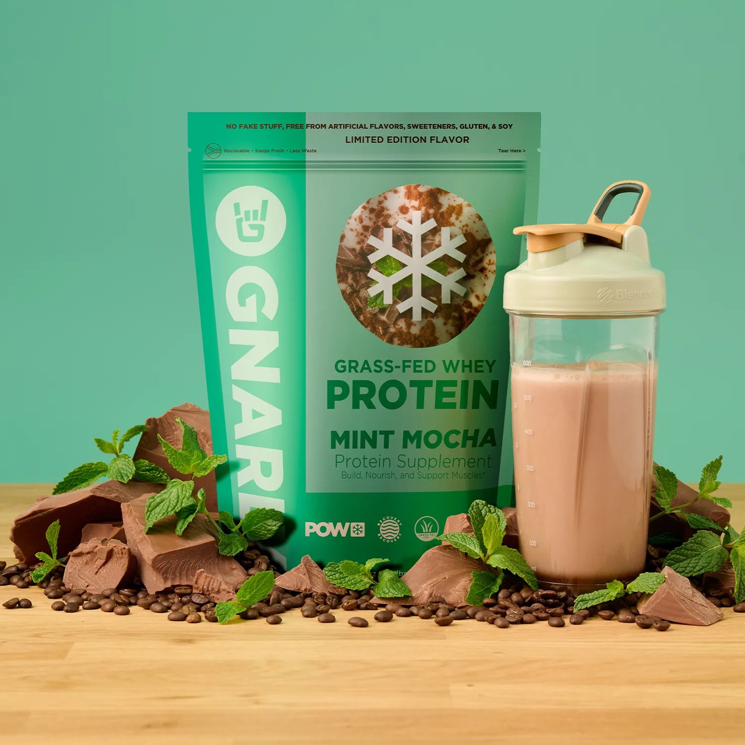 Green bag of Gnarly grass-fed whey protein mint mocha flavor with chocolate chunks, mint leaves, coffee beans, and a filled shaker bottle on a wooden surface.