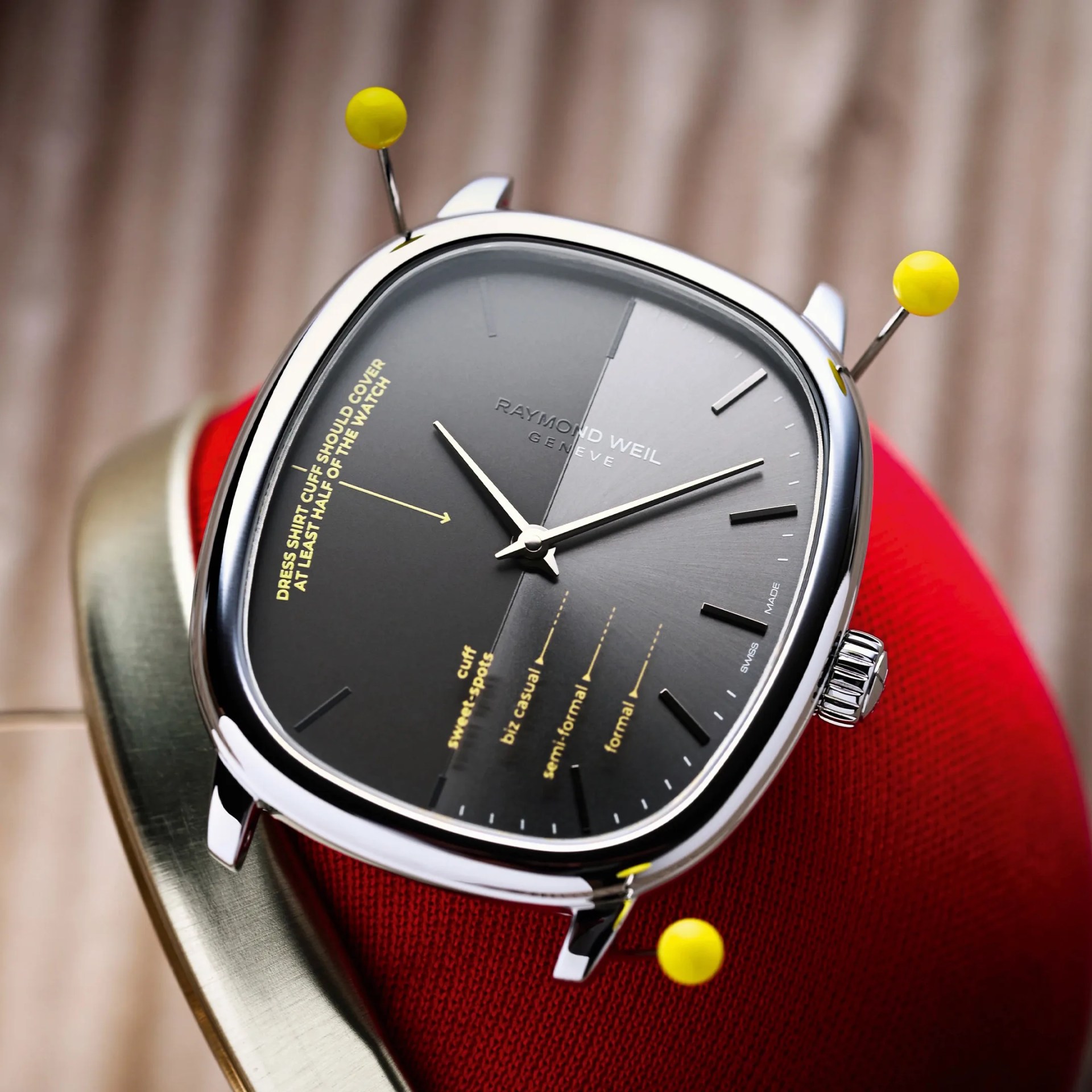 Square silver Raymond Weil wristwatch with black dial and yellow text, pinned on red fabric.