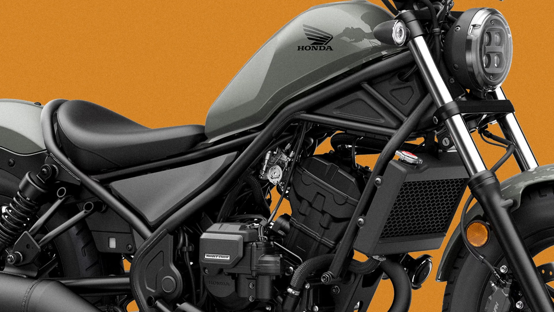 Close-up of a matte green Honda motorcycle fuel tank and black engine with frame and front suspension against an orange background.