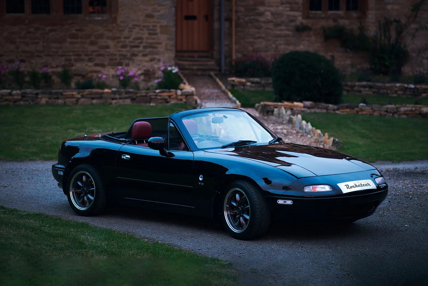 Black convertible sports car with red interior parked on a gravel driveway in front of a stone building.