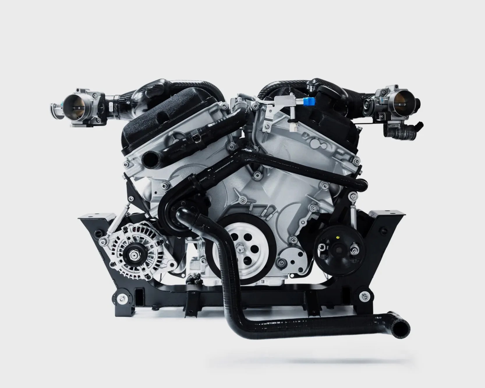 V-shaped car engine with black hoses and silver components mounted on a black frame against a white background.
