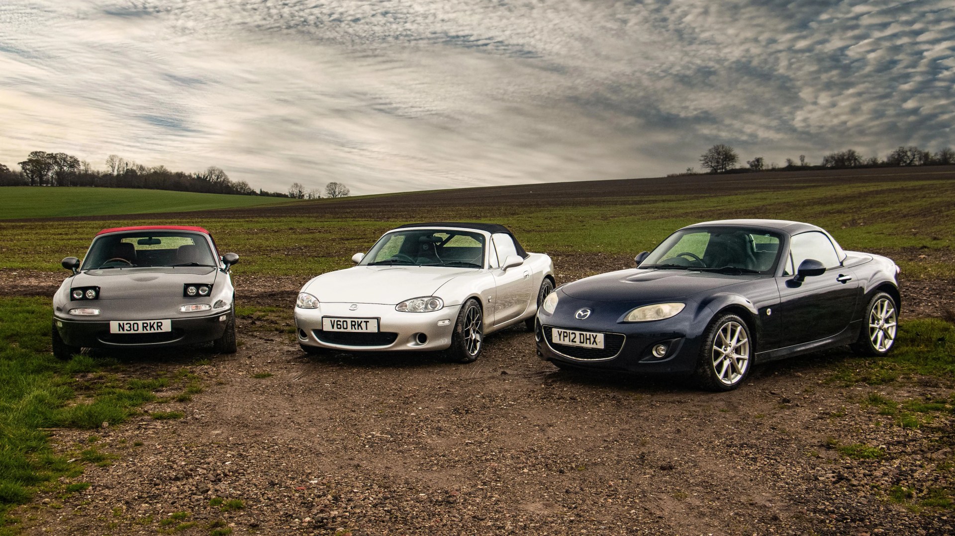 Three Mazda MX-5 Miata cars in black, silver, and dark blue parked on a dirt path with grassy fields and a cloudy sky in the background.