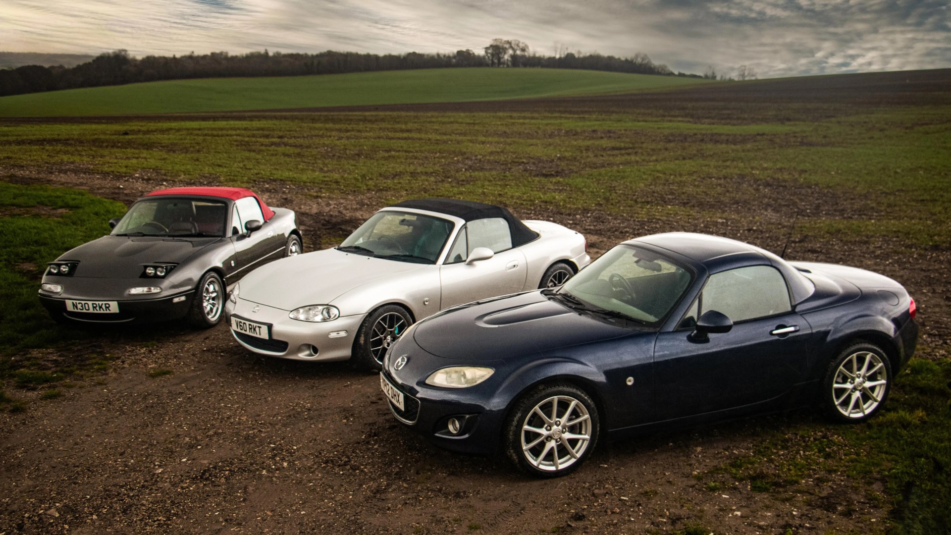 Three Mazda MX-5 Miata convertibles in black with red soft top, silver with black soft top, and dark blue with hardtop, parked on a dirt path near a green field.