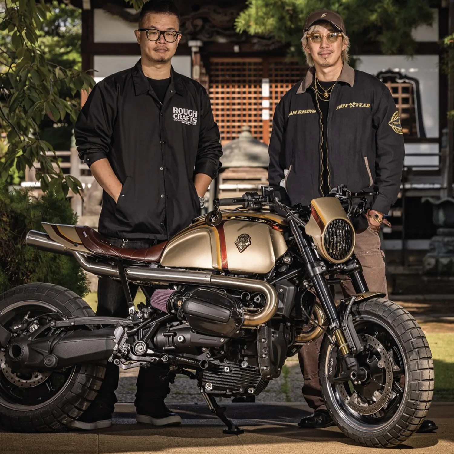 Two men standing behind a custom gold and brown motorcycle with thick tires in an outdoor setting.
