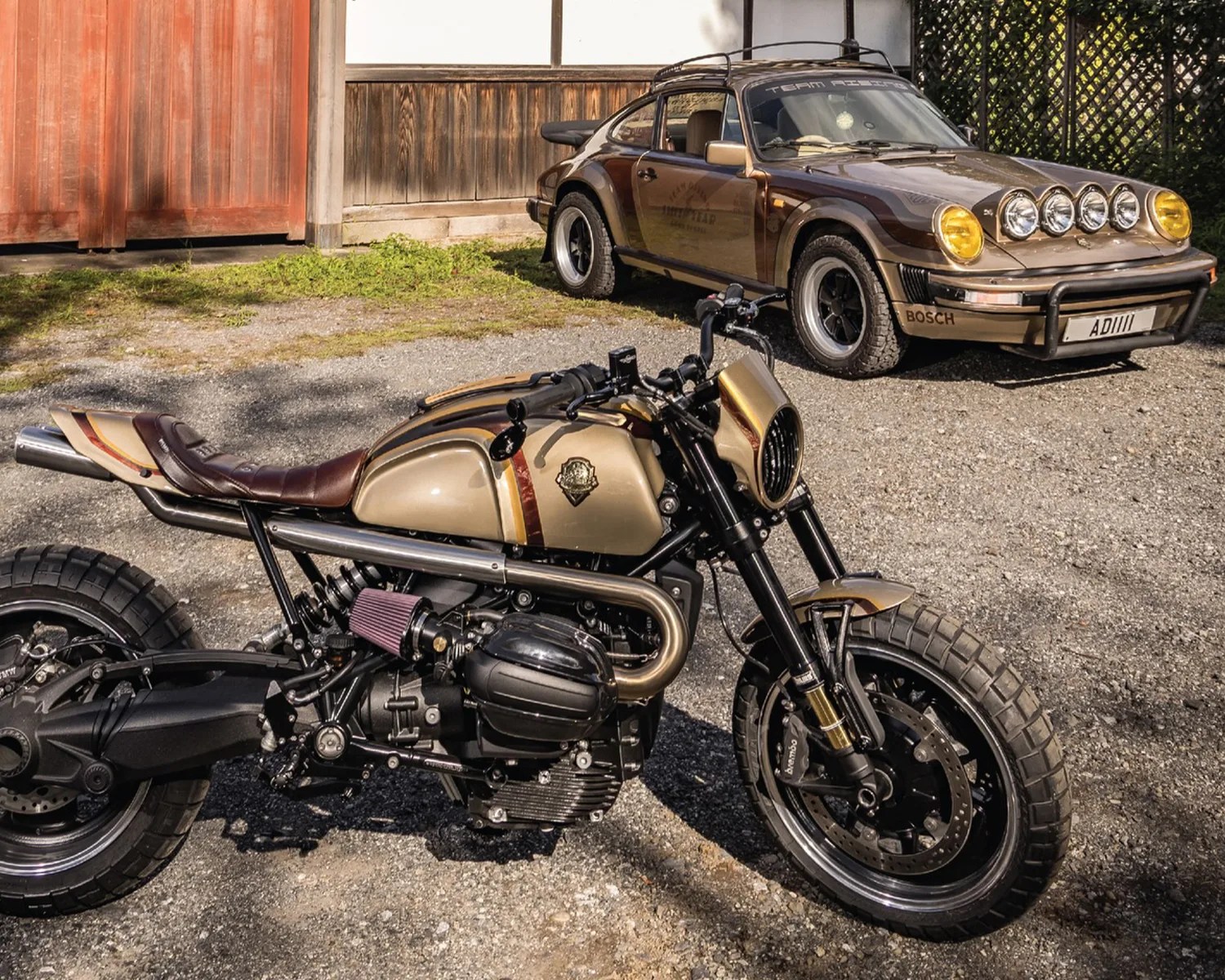 Custom beige and brown motorcycle with a leather seat parked on gravel, with a vintage brown car in the background.