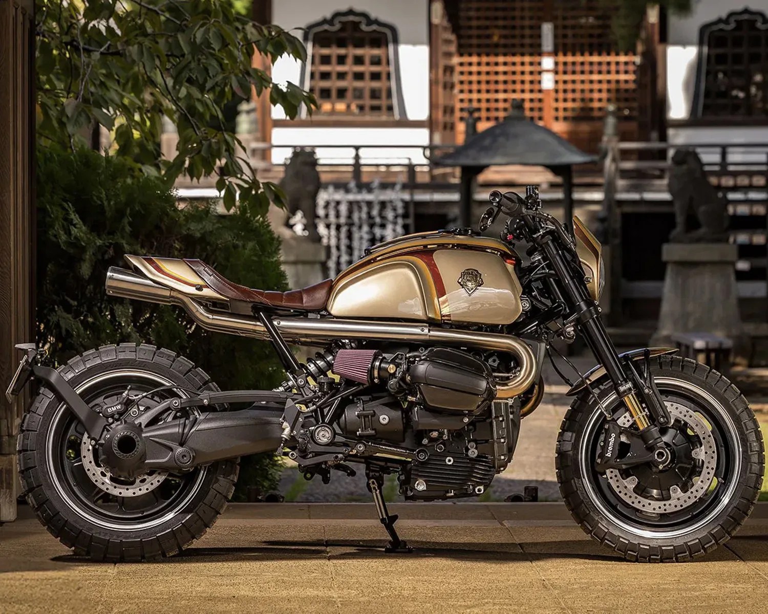 Custom gold and brown cafe racer motorcycle with knobby tires parked on a stone floor in front of a traditional wooden building.