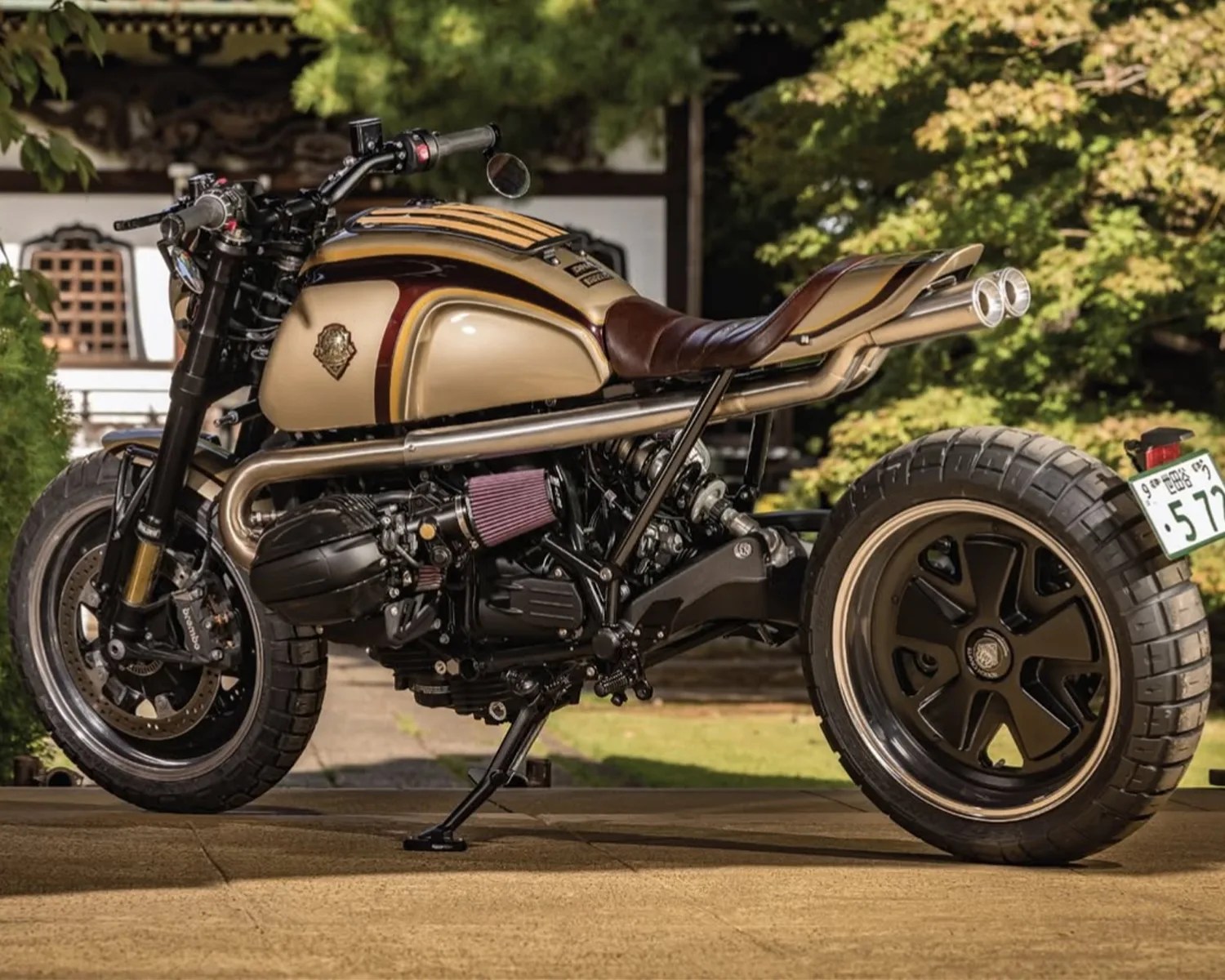 Custom beige and brown cafe racer motorcycle with large rear tire and dual exhaust pipes.