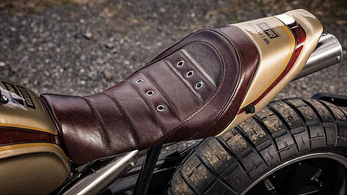 Brown leather motorcycle seat with metal eyelets on a gold and red rear fender above a knobby tire.