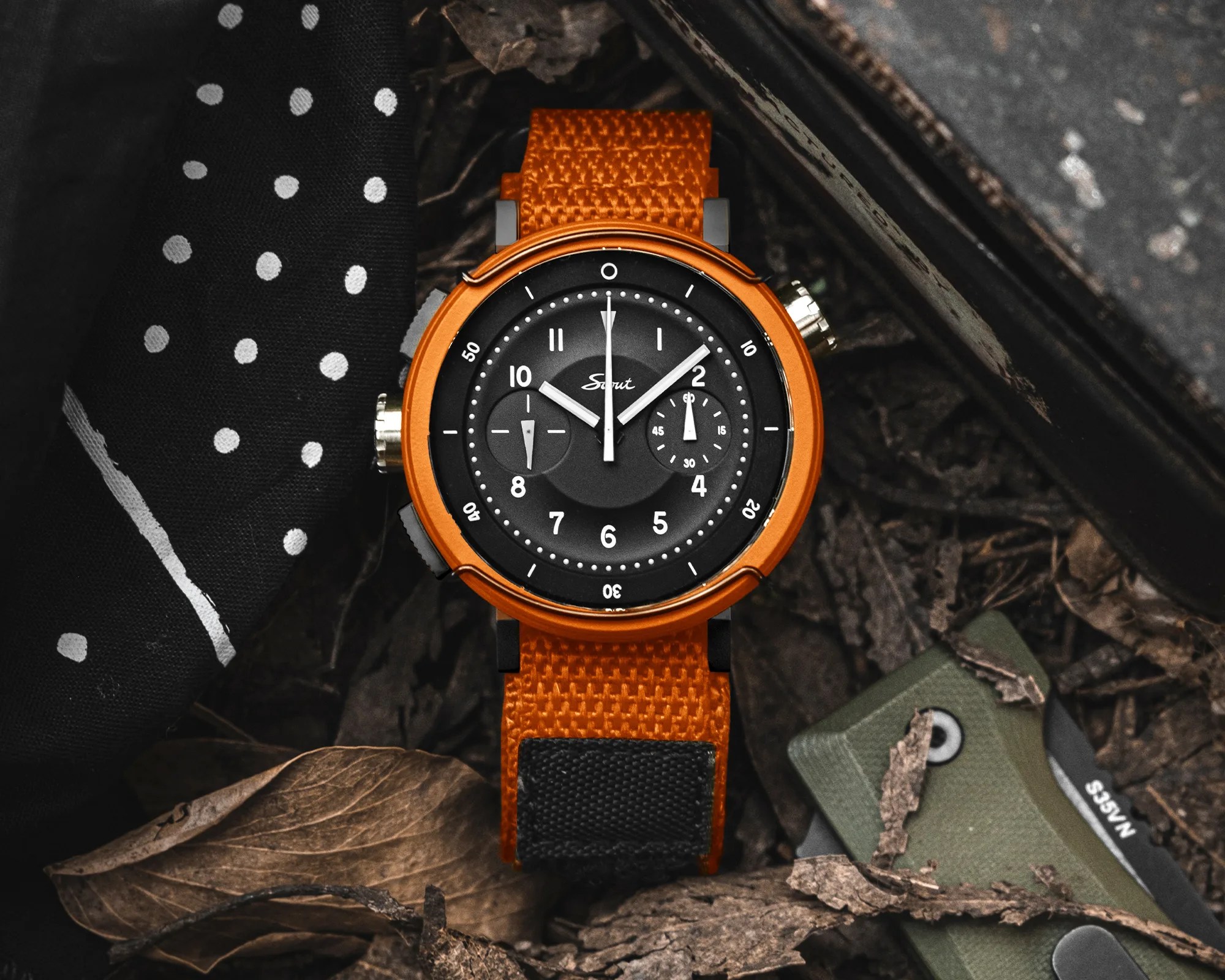 Orange wristwatch with black dial and orange fabric strap placed on dry leaves next to a black polka dot fabric.