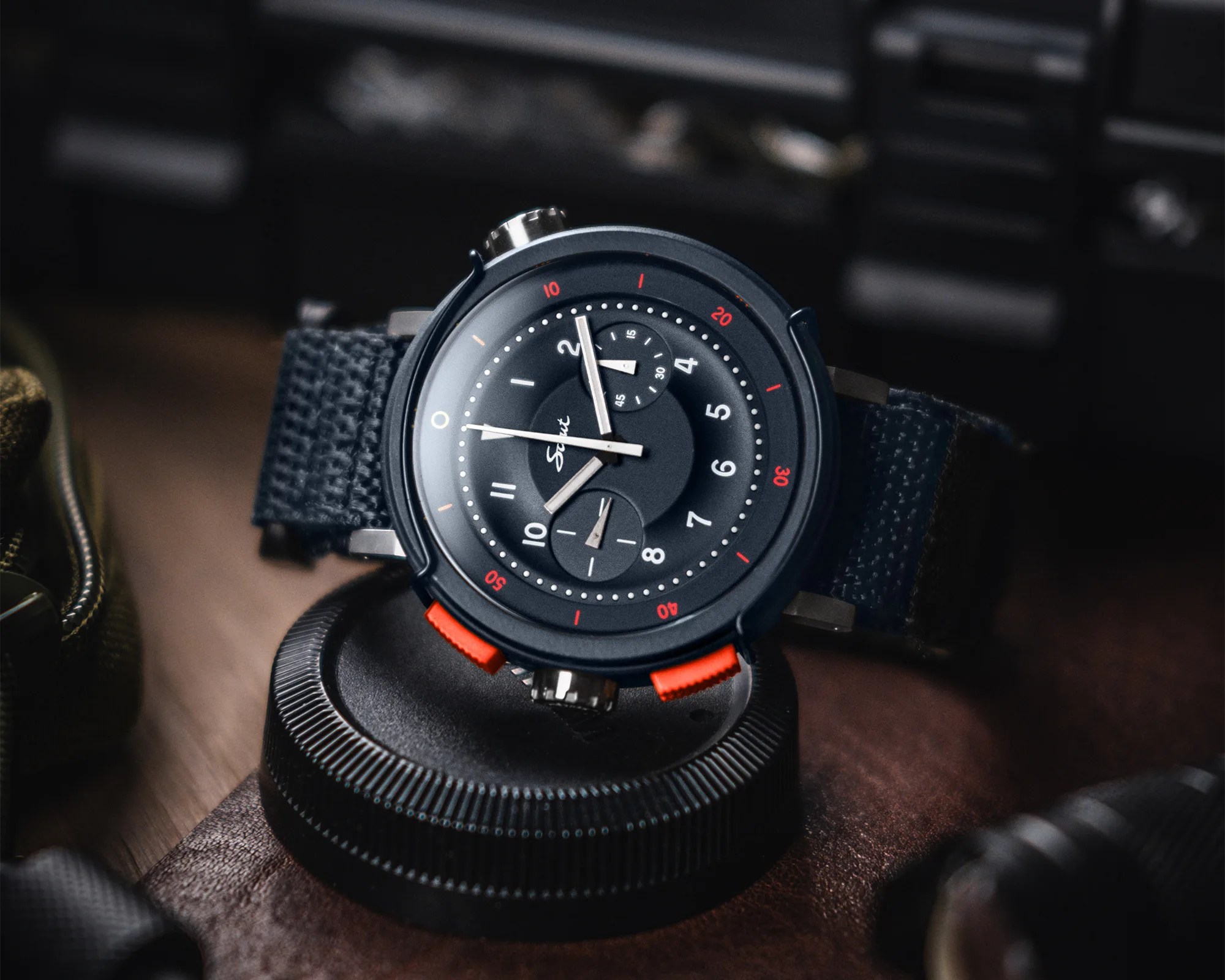 Black wristwatch with red accents and fabric strap resting on a textured black circular object.