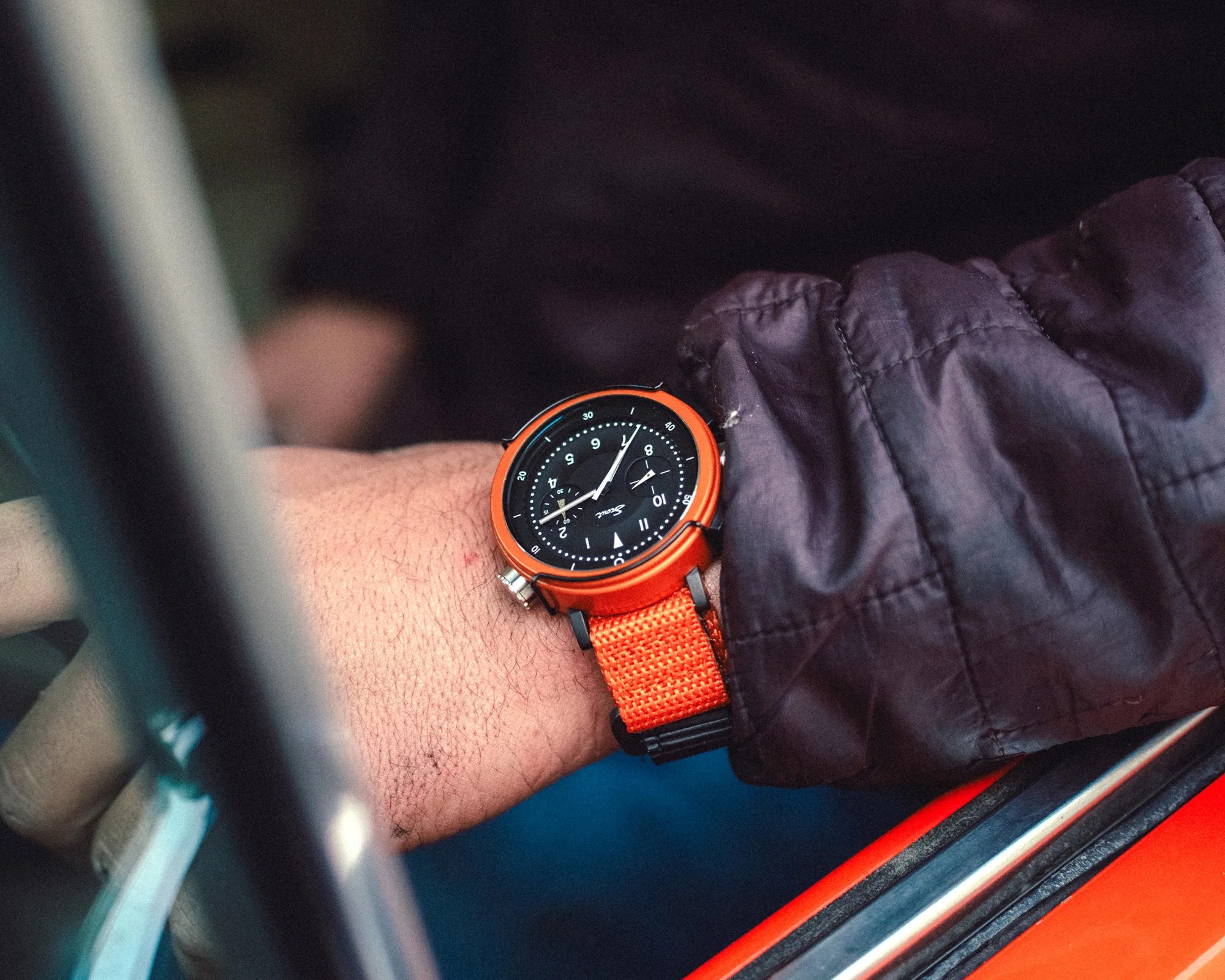Black dial wristwatch with orange bezel and orange fabric strap worn on a wrist with a dark jacket sleeve.