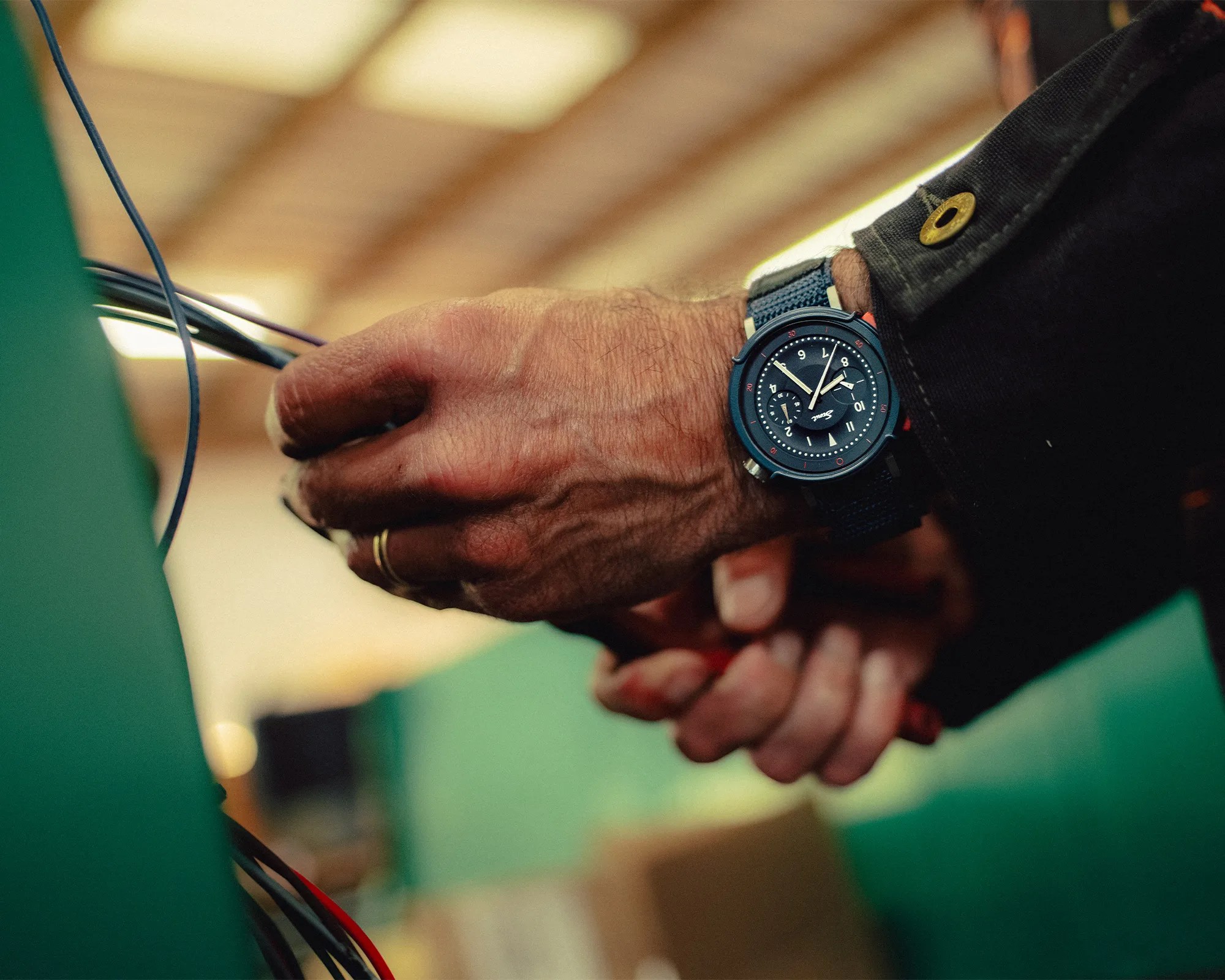 Black wristwatch with a blue fabric strap worn on a hand holding black and red wires.