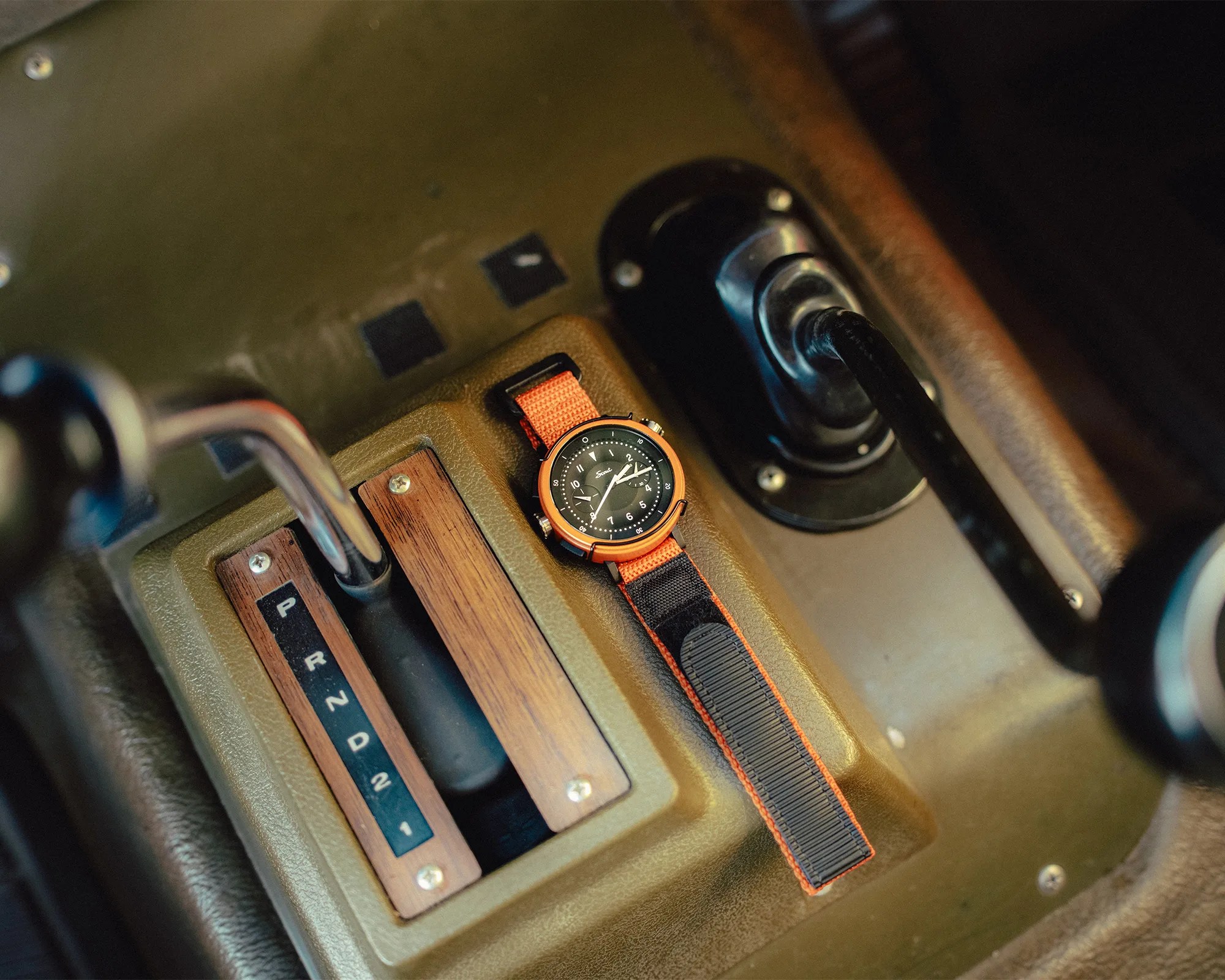 Black watch with orange bezel and orange and black fabric strap placed on a car's gear shift panel.