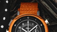 Close-up of a black watch face with white markings and hands, orange bezel, and textured orange strap.