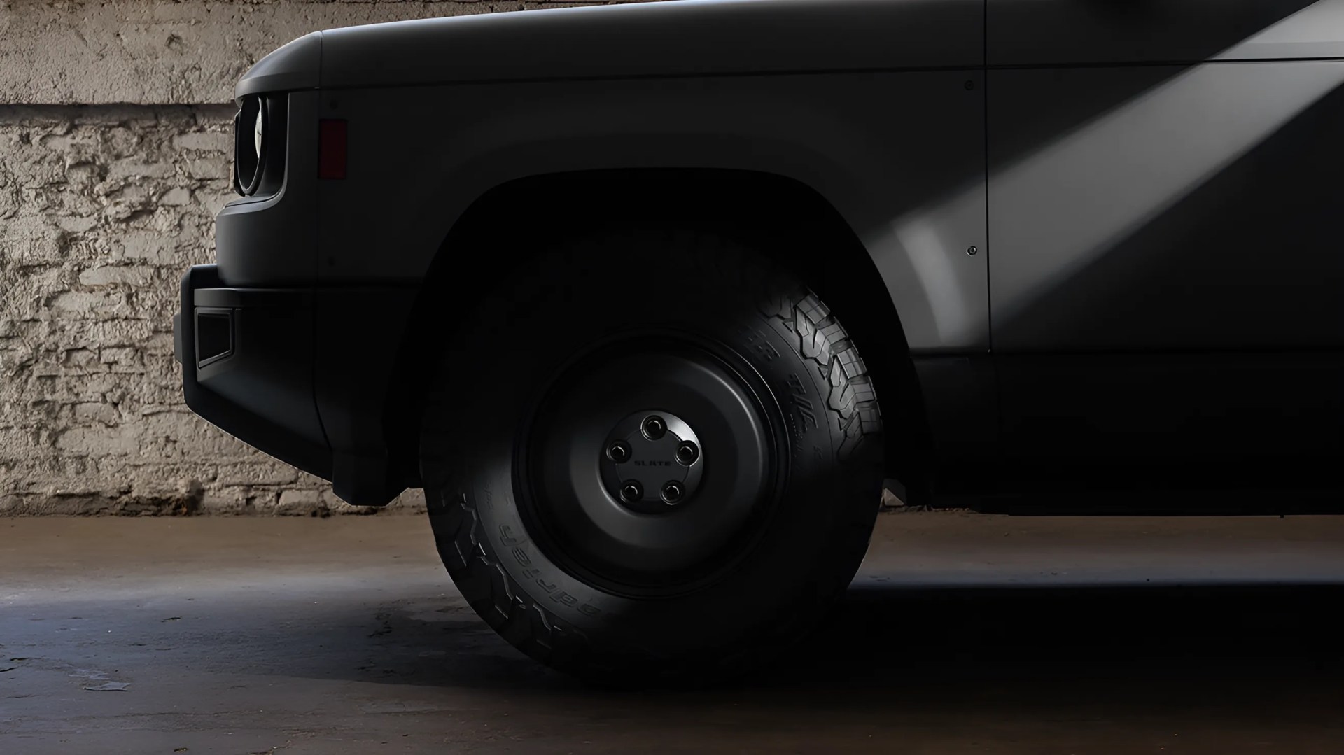 Close-up of a black off-road tire with a matte black rim on a dark gray vehicle in a dimly lit garage.