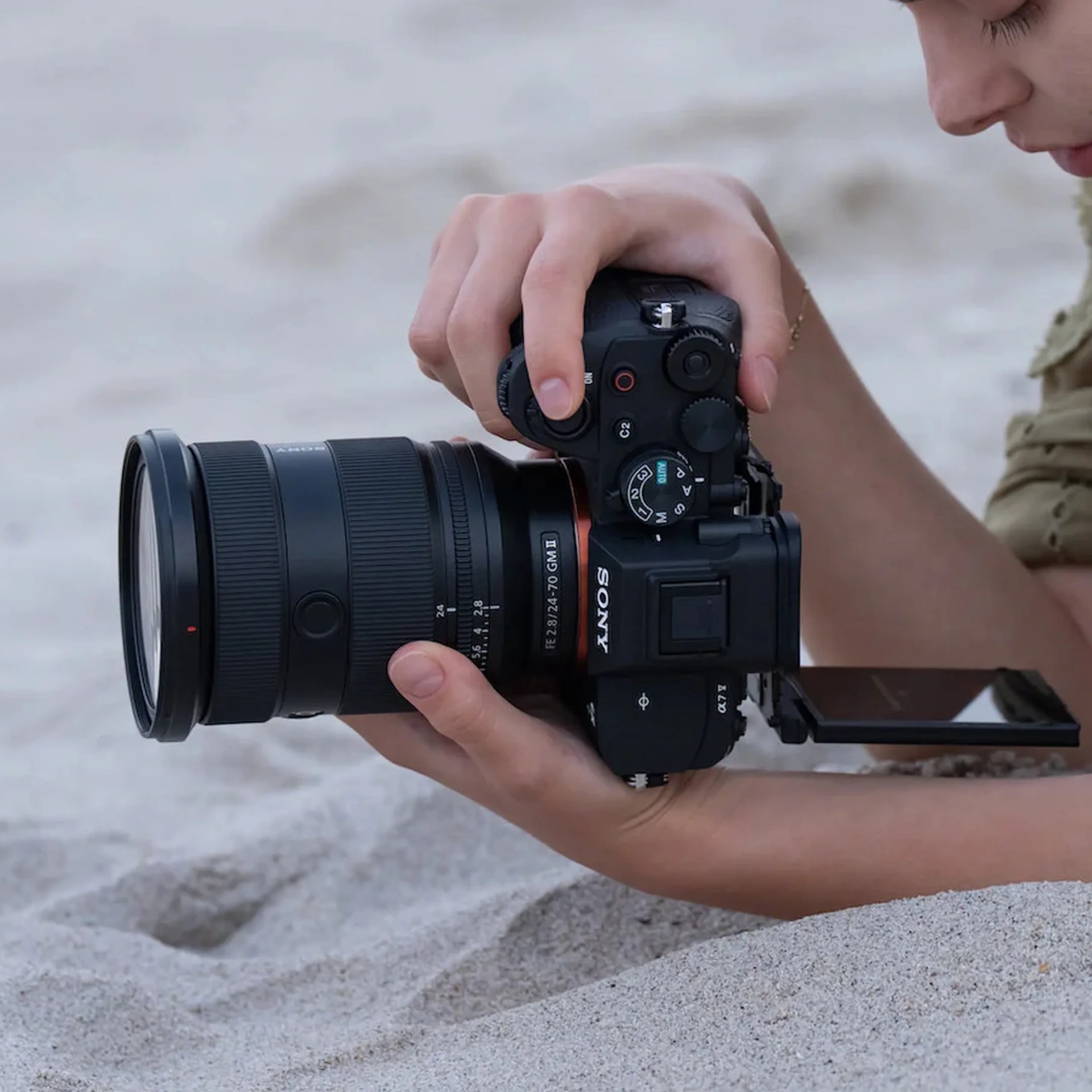 Person holding a Sony Alpha 7 IV camera with a 24-70mm GM II lens on sandy ground.