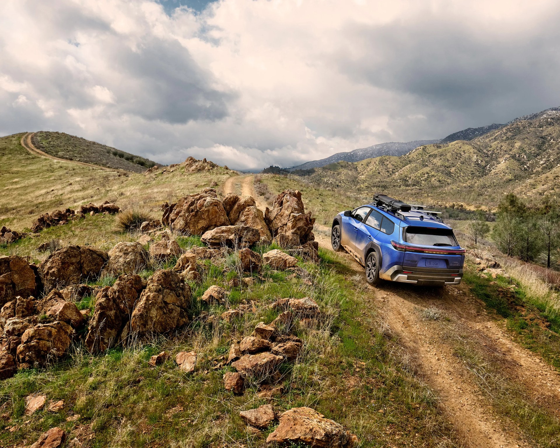 Blue SUV with roof rack driving on a dirt trail in a rocky, grassy mountainous landscape under cloudy sky.