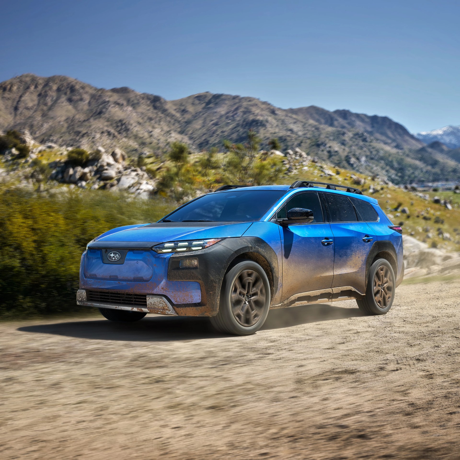 Blue Subaru SUV with mud splatters driving on a dirt road with mountains in the background.