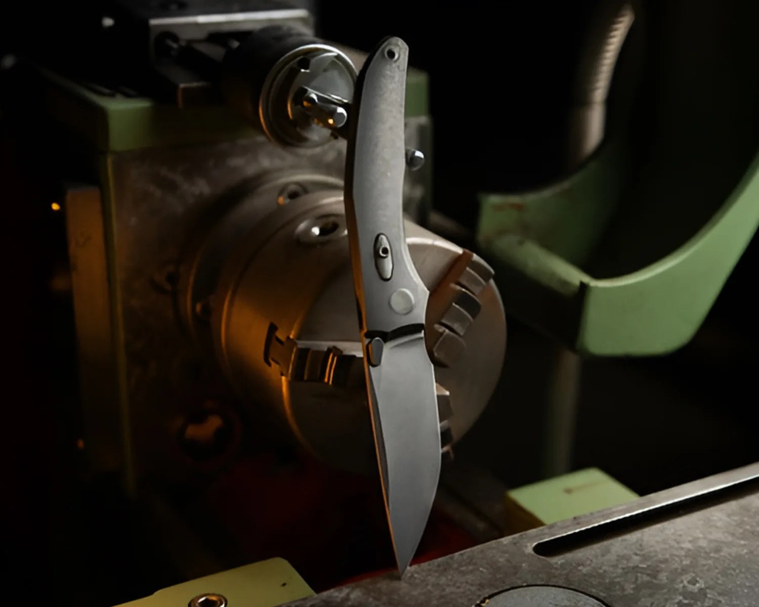Matte gray folding knife with a clip and thumb stud, positioned vertically on a metal lathe machine.