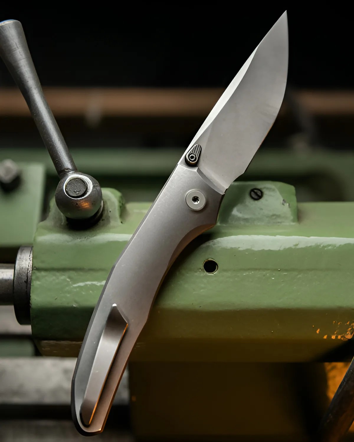 Silver folding knife with a clip resting on a green metal machine part.