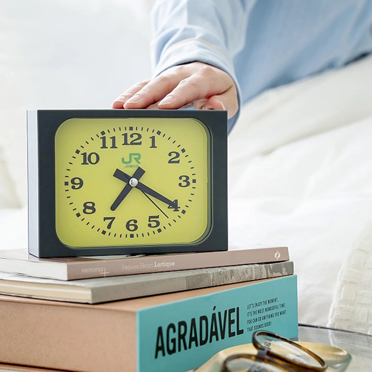Black-framed yellow analog clock showing 7:20 on a stack of books with a hand reaching to press it.