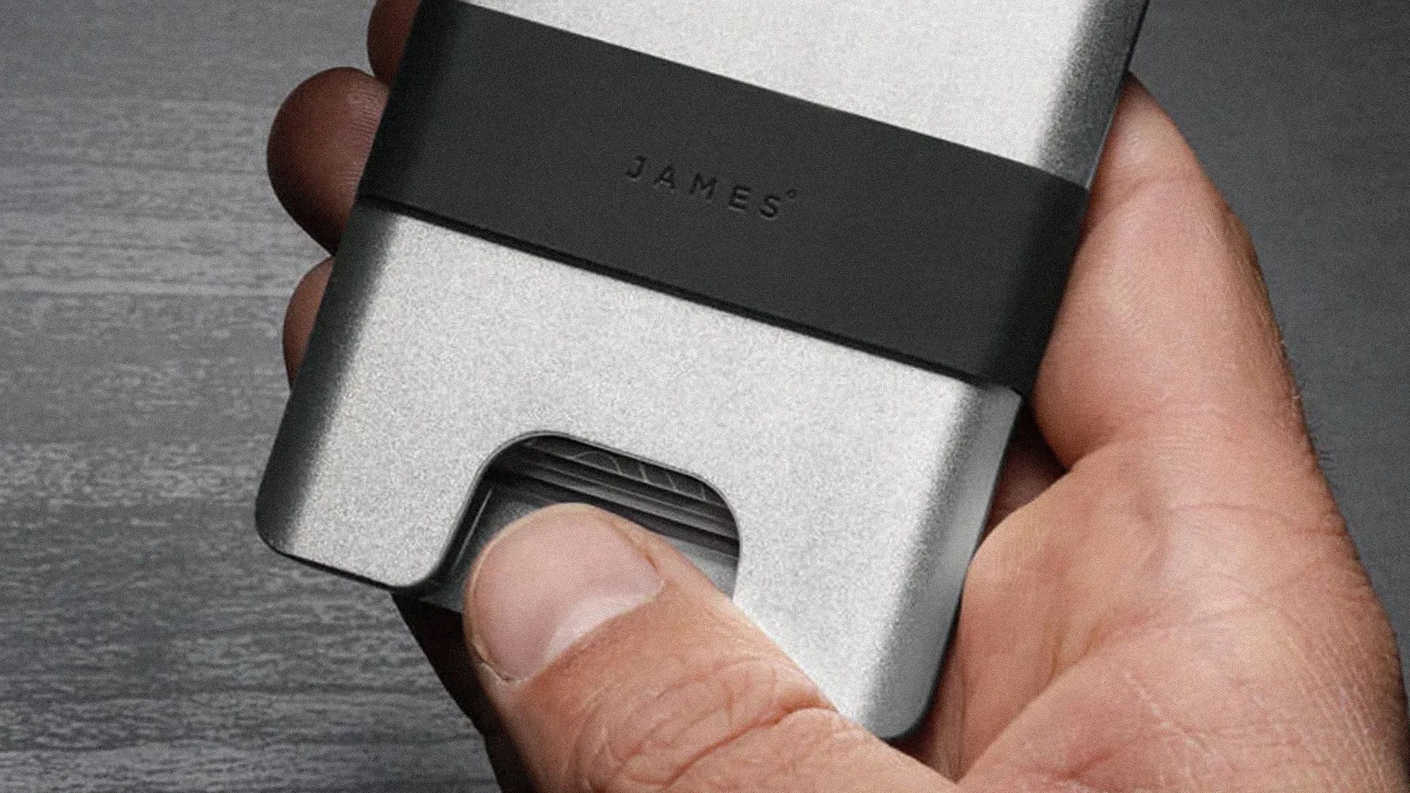 Hand holding a silver metal wallet with a black band labeled "JAMES" and cards partially visible.