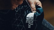 Close-up of a hand sewing white thread on a black fabric with The North Face logo and white stitched geometric patterns.