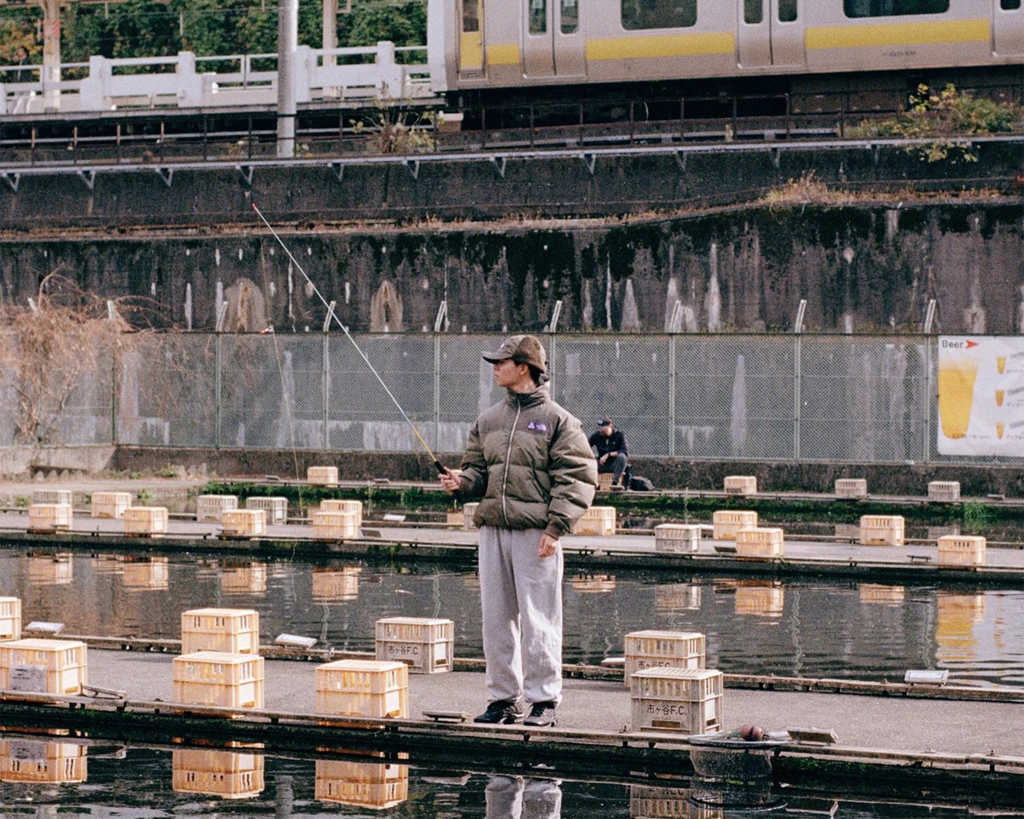 Person in a green puffer jacket and cap fishing on a dock with yellow crates and a train passing in the background.