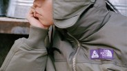 Close-up of a person wearing an olive-green jacket with purple Palace and The North Face logos on the chest.