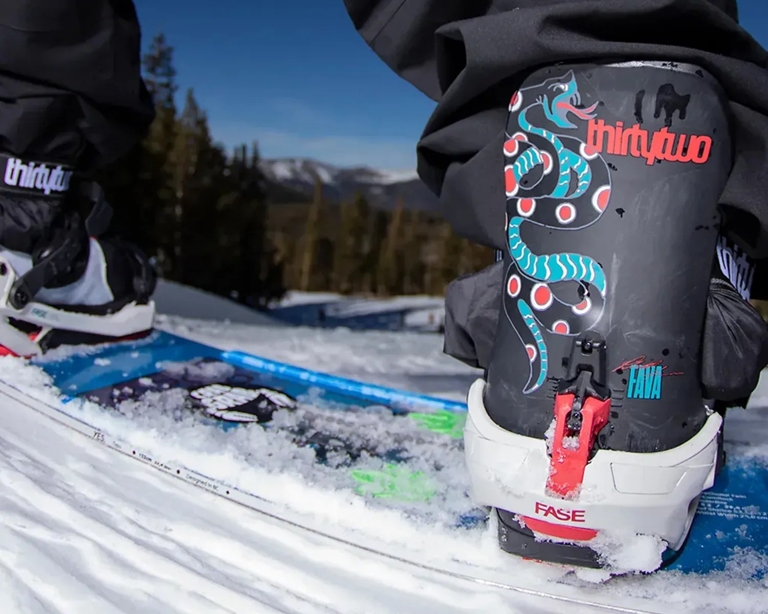 Close-up of black ski boots with colorful snake design and red-white bindings on a snow-covered ski.