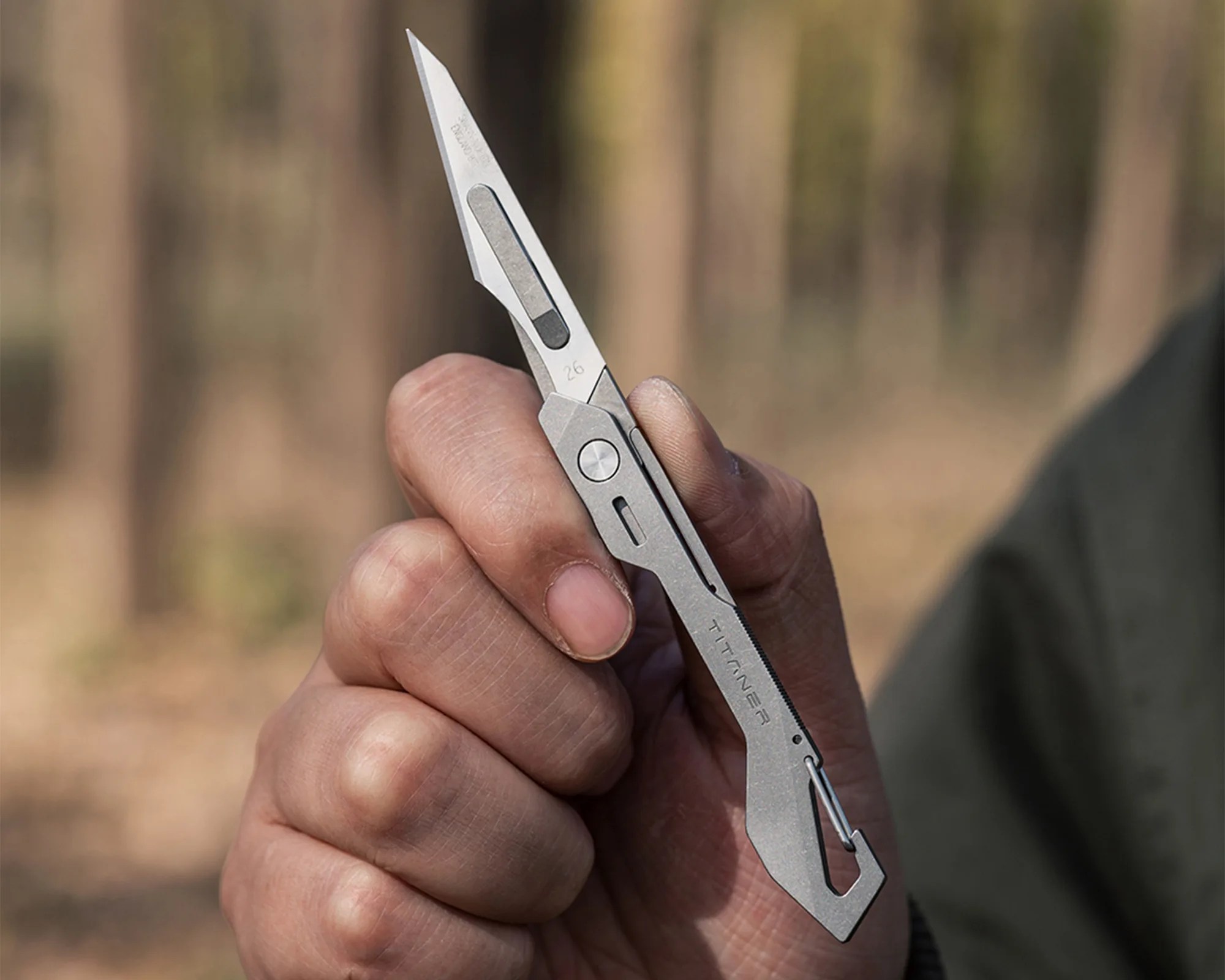 Hand holding a slim, silver titanium folding knife with a pointed blade and integrated carabiner clip.