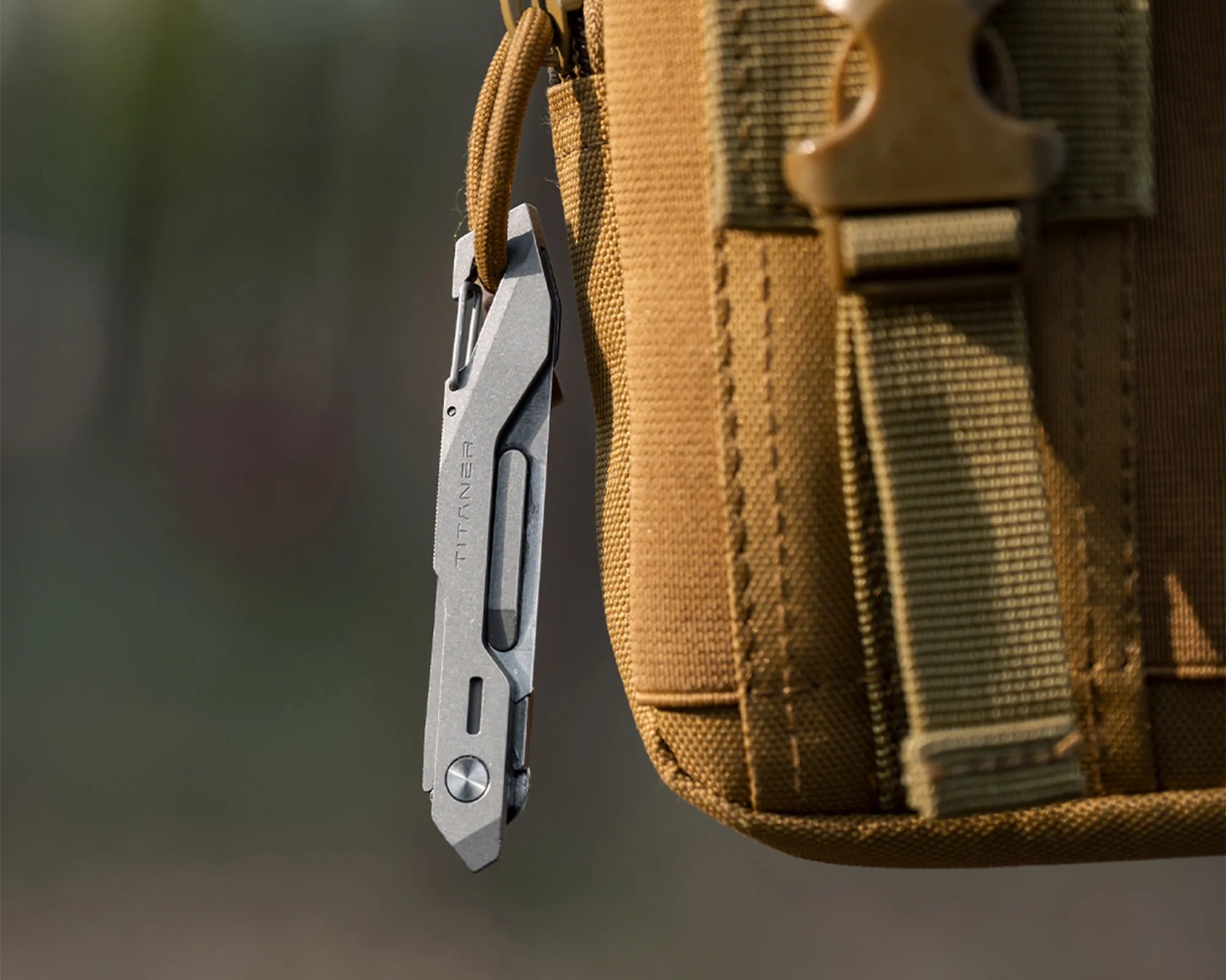 Gray metal multitool with "TITANER" engraving attached to a tan fabric bag with webbing straps.