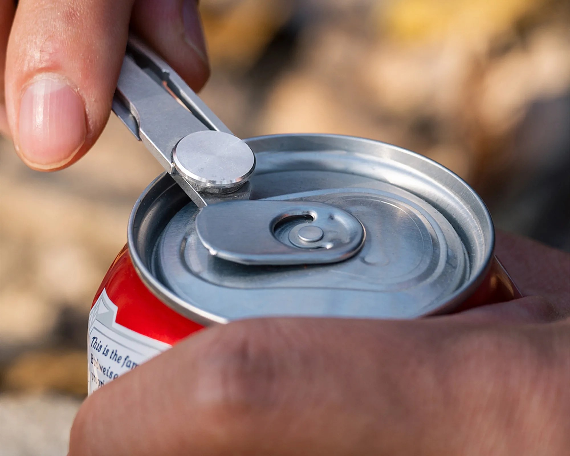 Hand using a metal tool to open the tab of a red aluminum beverage can.