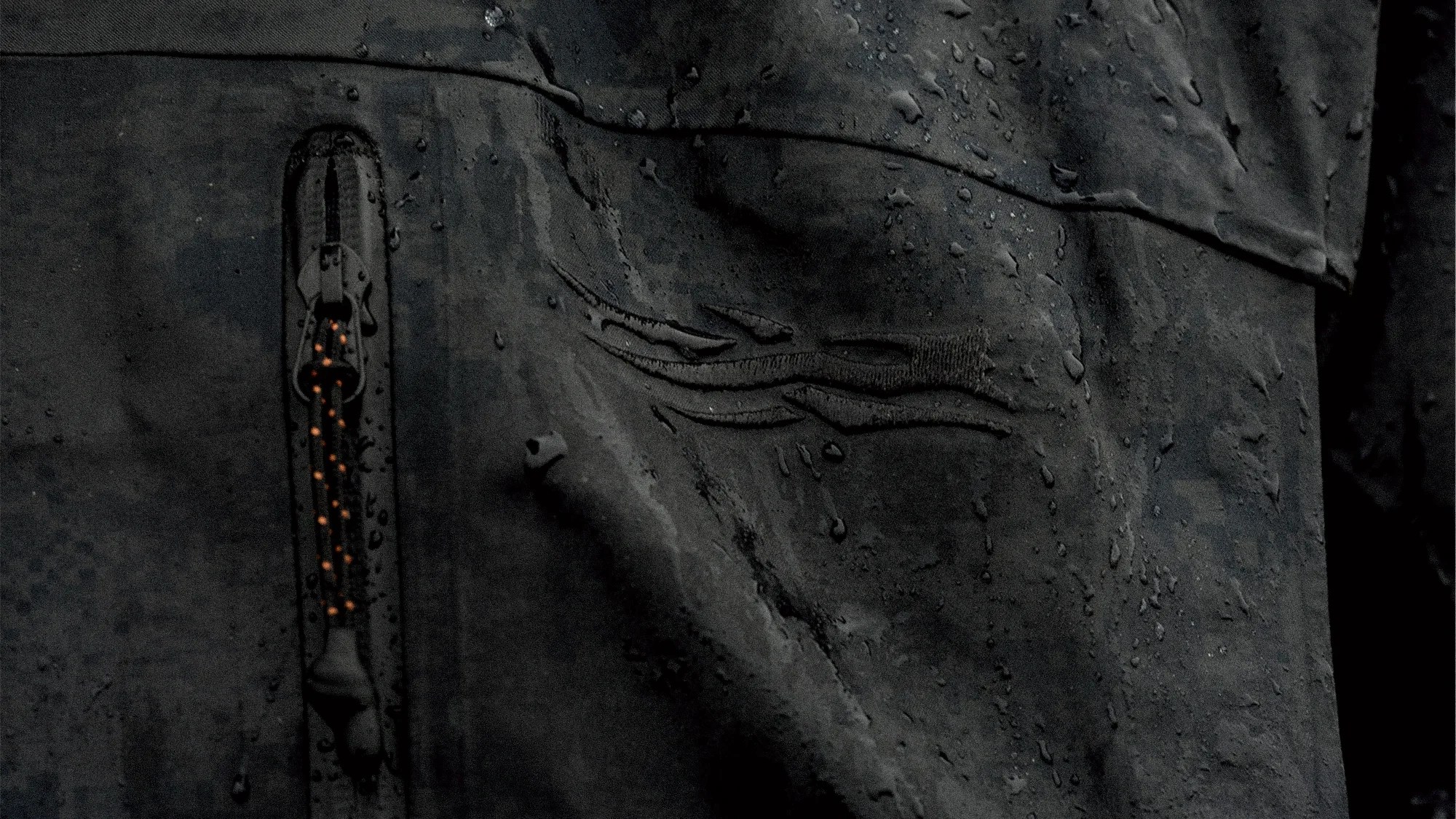 Close-up of a black waterproof jacket with water droplets on the fabric and a zipper with an orange pull cord.