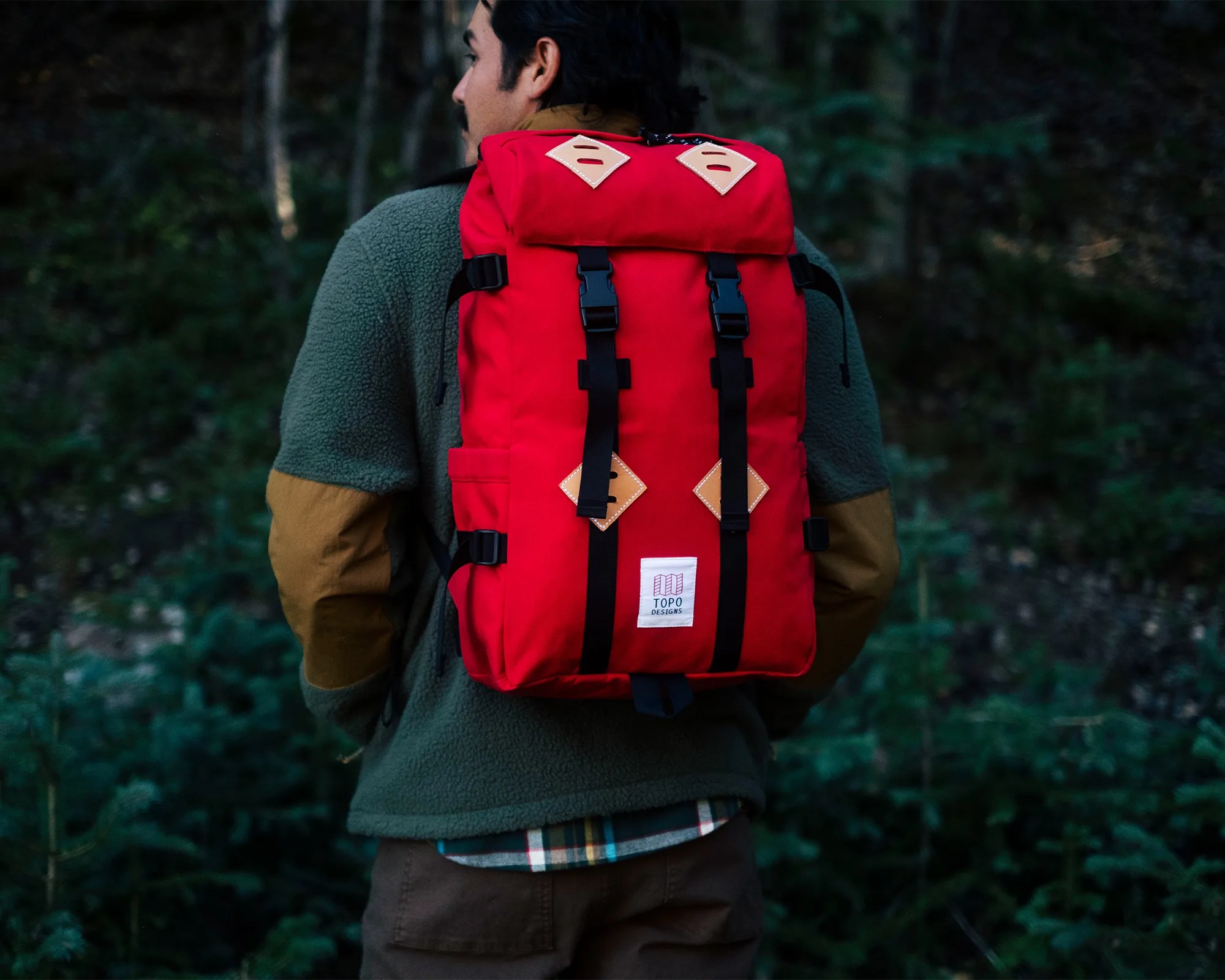 Red Topo Designs backpack with black straps and tan leather accents worn by a person in a green fleece jacket in a forest.