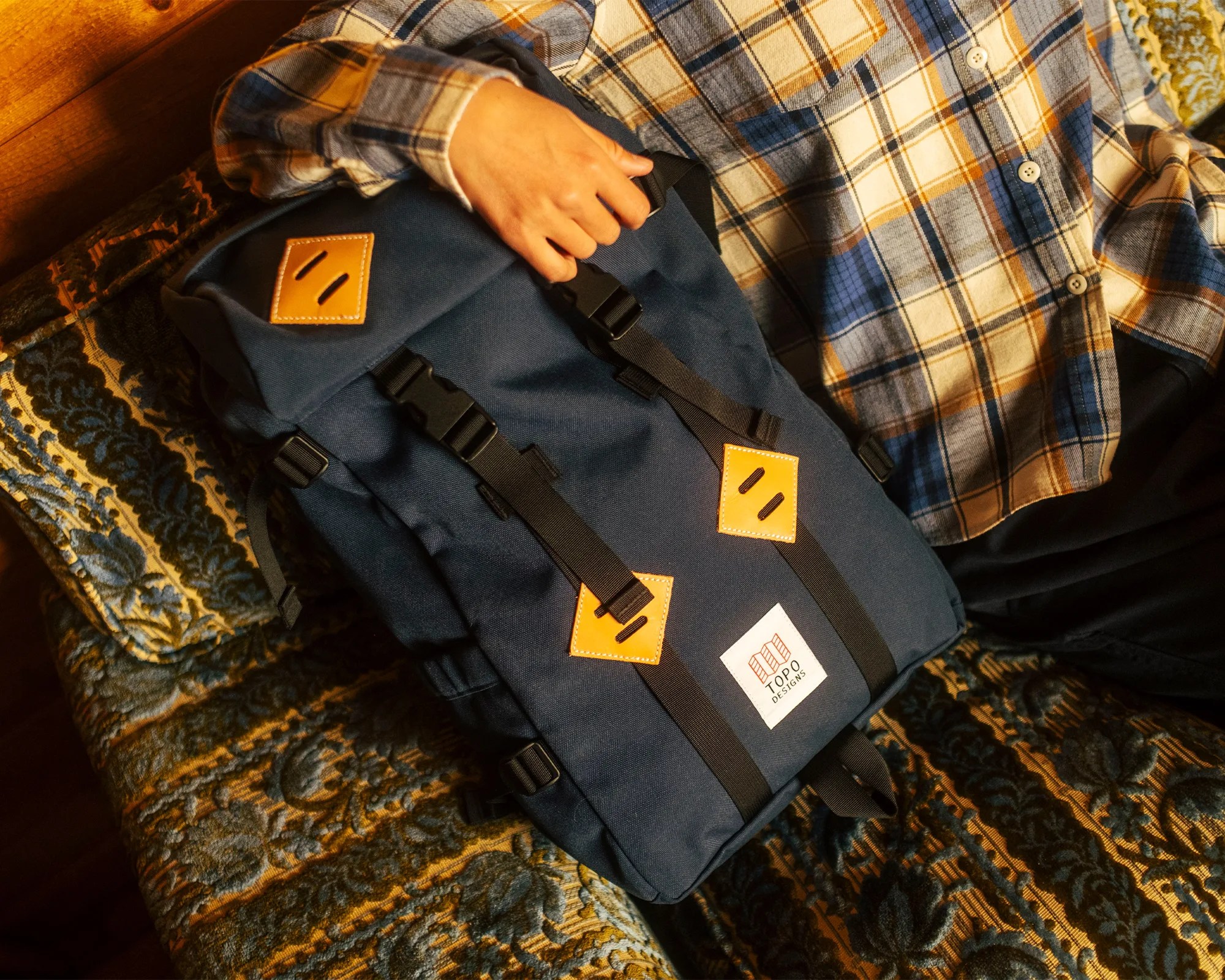 Navy blue Topo Designs backpack with black straps and tan leather patches held by a person wearing a plaid shirt.