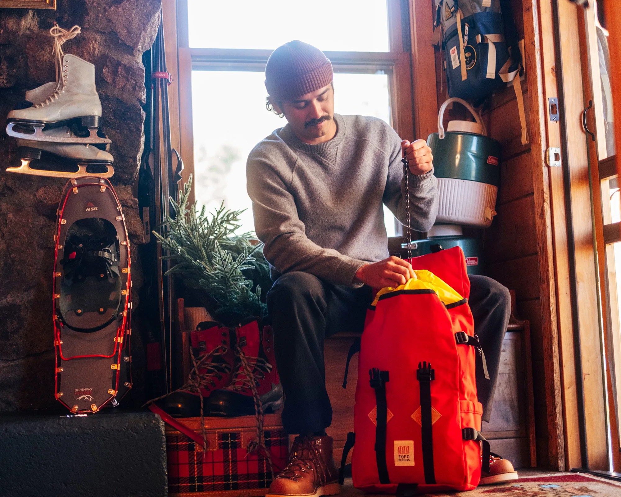 Person sitting indoors near a window, wearing a gray sweater and brown boots, packing a bright red Topo Designs backpack.